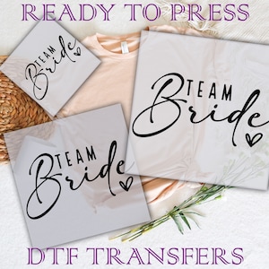 May include: Three white iron-on transfers with the text "Team Bride" in black script with a heart. The transfers are ready to press onto clothing.
