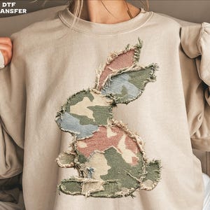 May include: Beige sweatshirt featuring a distressed camouflage bunny design. The bunny is made from patches of pink, blue, and green camouflage fabric. The text "DTF TRANSFER" is visible in the upper left corner.
