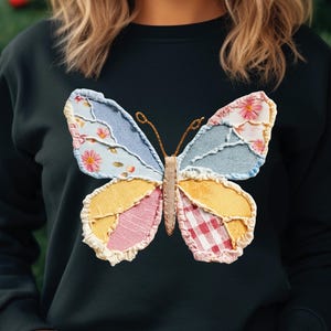 May include: Black sweatshirt featuring a colorful butterfly applique. The butterfly design incorporates floral, gingham, and denim-like fabrics in shades of blue, pink, yellow, and red. The butterfly's body and antennae are a tan color.