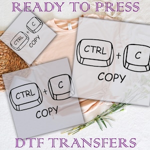May include: Three sheets of clear transfer paper with black and white keyboard key graphics that say "CTRL + C" and "COPY". The sheets are on top of a light brown t-shirt.