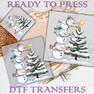 May include: Three snowmen stacked on top of each other, wearing hats and scarves, stand next to a green Christmas tree with white snow on the branches. The snowmen are holding a star. The image is on a white background with the text "READY TO PRESS" at the top and "DTG TRANSFERS" at the bottom.