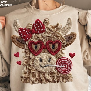 May include: Beige sweatshirt featuring an embroidered Highland cow design. The cow has heart-shaped sunglasses, a red bow with white polka dots, and a lollipop. Small red hearts are scattered around the cow.