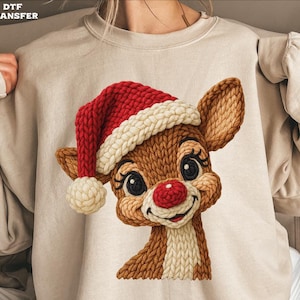 May include: A beige sweatshirt featuring a knitted Rudolph the Red-Nosed Reindeer design. The reindeer wears a red and white Santa hat and has a red nose. The design is detailed with brown, beige, and white knitted textures.