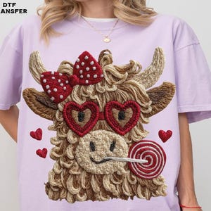 Pode incluir: Camiseta lil&aacute;s claro com um design bordado de uma vaca das Terras Altas com &oacute;culos de sol em forma de cora&ccedil;&atilde;o, um la&ccedil;o vermelho e um pirulito. O design inclui pequenos cora&ccedil;&otilde;es vermelhos. O texto "DTF TRANSFER" &eacute; vis&iacute;vel.