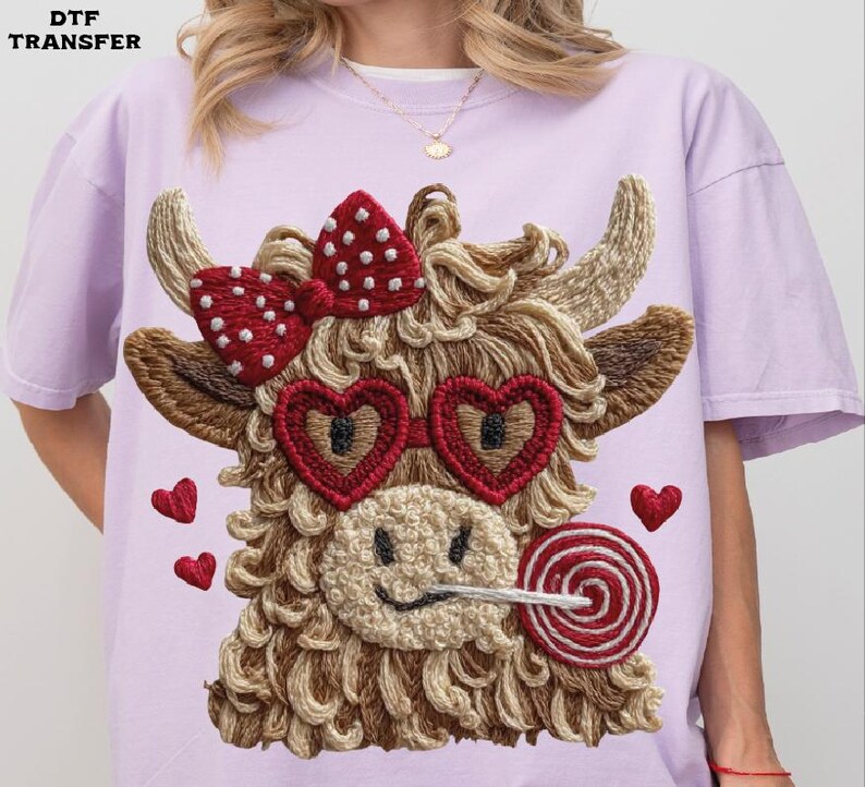 Pode incluir: Camiseta lil&aacute;s claro com um design bordado de uma vaca das Terras Altas com &oacute;culos de sol em forma de cora&ccedil;&atilde;o, um la&ccedil;o vermelho e um pirulito. O design inclui pequenos cora&ccedil;&otilde;es vermelhos. O texto "DTF TRANSFER" &eacute; vis&iacute;vel.