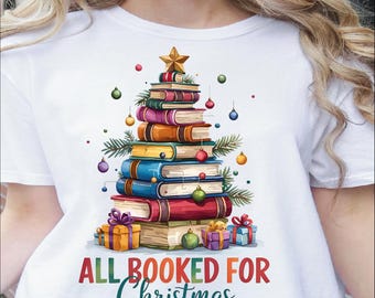 May include: White t-shirt featuring a colorful illustration of a Christmas tree made of stacked books. The design includes ornaments, a star, and the text "ALL BOOKED FOR Christmas".
