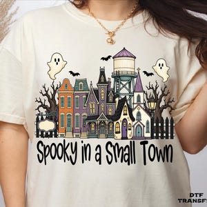 May include: A cream-colored t-shirt featuring a graphic of a small town with colorful houses, a water tower, bare trees, bats, and two ghosts. The text "Spooky in a Small Town" is printed in black below the illustration, with "DTF TRANSFER" in the corner.