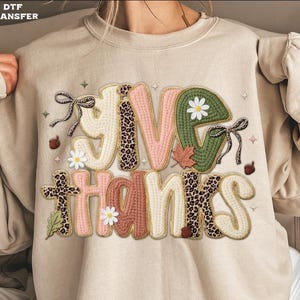May include: Beige sweatshirt with the words "GIVE THANKS" in a decorative, stitched design. The letters feature a mix of textures, including leopard print, pink, green, and cream, with floral and leaf accents. A fall-themed apparel item.