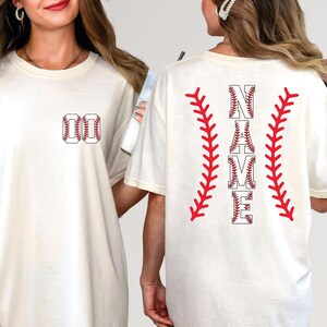 May include: Cream-colored t-shirt with baseball-themed design. The front features the number "00" in a baseball stitch design. The back displays the word "NAME" in a baseball font, flanked by red baseball stitching.