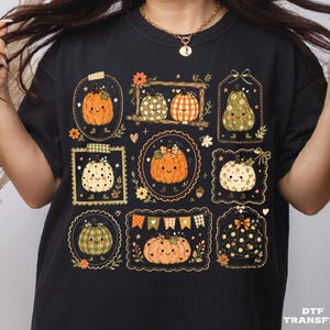 May include: A black t-shirt featuring a graphic print of multiple smiling pumpkins in various patterns and colors, including orange, green, and white, arranged within decorative frames, tags, and borders with small floral and bunting accents.