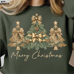 May include: Green sweatshirt with a Christmas tree design. The design features three Christmas trees with leopard print bows and gold accents. The text "Merry Christmas" is written in a cursive font.