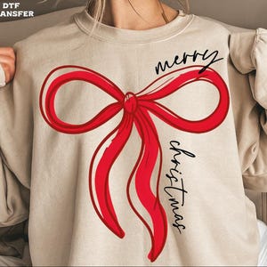 May include: Beige sweatshirt featuring a large red bow design with the words "merry christmas" in black script. The bow is outlined in red and has a hand-drawn aesthetic. A festive holiday-themed apparel item.