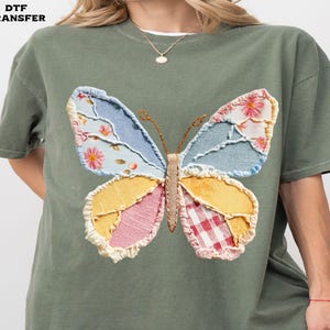May include: An olive green t-shirt featuring a large, colorful butterfly design. The butterfly has wings made of patchwork fabric in shades of blue, yellow, pink, and red. The text "DTF TRANSFER" is in the top left corner.