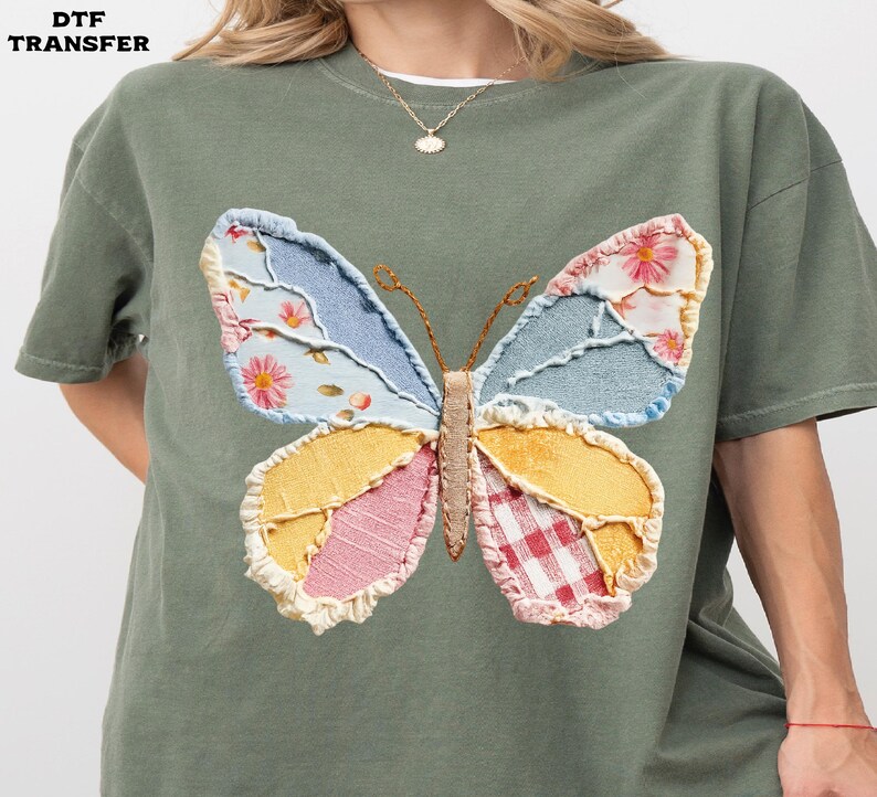 May include: An olive green t-shirt featuring a large, colorful butterfly design. The butterfly has wings made of patchwork fabric in shades of blue, yellow, pink, and red. The text "DTF TRANSFER" is in the top left corner.