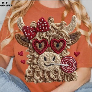 Pode incluir: Design bordado de uma vaca Highland usando &oacute;culos de sol em forma de cora&ccedil;&atilde;o e um la&ccedil;o de bolinhas. A vaca tem um pirulito na boca e pequenos detalhes de cora&ccedil;&atilde;o. O design est&aacute; em uma camiseta laranja.