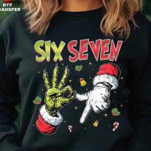 May include: Black sweatshirt with a graphic featuring a green hand making an 'okay' gesture and a white gloved hand pointing. The text 'SIX SEVEN' is in red and green. Festive holly and candy cane accents.