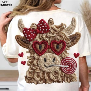 Pode incluir: Camiseta creme com um design bordado detalhado de uma vaca das Terras Altas com &oacute;culos de sol em forma de cora&ccedil;&atilde;o e um la&ccedil;o vermelho com bolinhas brancas. A vaca tamb&eacute;m segura um pirulito. Pequenos cora&ccedil;&otilde;es vermelhos est&atilde;o espalhados.