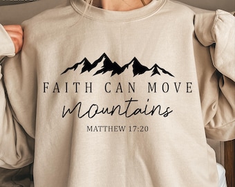 May include: Beige sweatshirt with the text "Faith Can Move Mountains" and a black mountain graphic. The text "Matthew 17:20" is below the main text. The sweatshirt is a crew neck style.