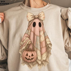 Peut inclure: Un sweat-shirt beige avec un motif de fantôme fantaisiste. Le fantôme est rose avec un motif patchwork, un nœud floral et un panier de citrouille. Le design a une esthétique vintage et cottagecore, parfait pour Halloween.