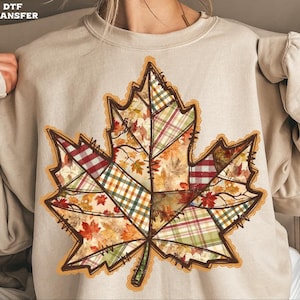 May include: Beige sweatshirt featuring a large, detailed maple leaf design. The leaf is composed of patchwork patterns in fall colors like red, green, and orange. The leaf has a brown outline and a decorative lace trim.