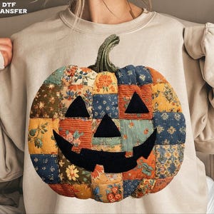 Peut inclure: Sweat-shirt beige avec un motif citrouille en patchwork. La citrouille est composée de divers carrés de tissu aux motifs floraux orange, bleu, jaune et rouge. Des triangles noirs forment les yeux et le nez, avec une bouche incurvée. La tige est verte.