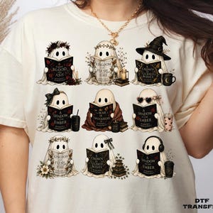 May include: A cream-colored t-shirt featuring a graphic print of nine cartoon ghosts reading books. Each ghost is depicted with different accessories like hats, flowers, or glasses, and the books display titles by Jennifer L. Armentrout.