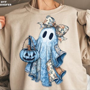 May include: A beige sweatshirt featuring a whimsical ghost design. The ghost is light blue with patches of floral fabric, a floral bow, and a blue pumpkin basket. The design is a DTF transfer.