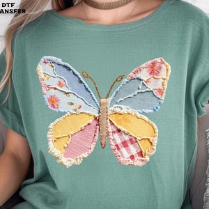 May include: A light green t-shirt featuring a colorful butterfly design. The butterfly has wings made of patchwork fabric in various patterns and colors, including floral, denim, and gingham. The text "DTF TRANSFER" is in the upper left corner.