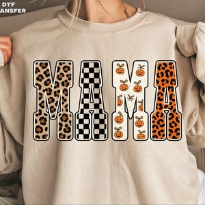 May include: Beige sweatshirt featuring the word "MAMA" in a decorative design. The letters are patterned with leopard print, black and white checks, pumpkins, and orange leopard print. The design has an autumnal theme.