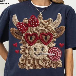 Pode incluir: Camiseta azul marinho com um design bordado de vaca Highland. A vaca tem &oacute;culos de sol em forma de cora&ccedil;&atilde;o, um la&ccedil;o vermelho e um pirulito. Pequenos cora&ccedil;&otilde;es vermelhos est&atilde;o espalhados. A camiseta tem gola redonda.