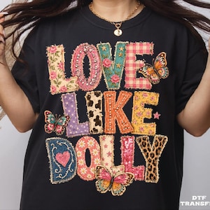 May include: A black t-shirt featuring a multi-patterned graphic print that reads LOVE LIKE DOLLY. The letters are decorated with floral, gingham, cow print, leopard print, and sequin textures, accented by three colorful butterfly illustrations.