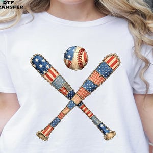 May include: White t-shirt featuring a baseball and two crossed baseball bats. The baseball and bats are decorated with a patchwork design of the American flag. The bats are brown with red, white, and blue patches.