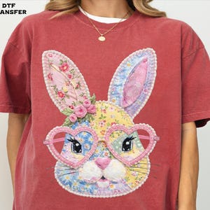 May include: Red t-shirt featuring a colorful bunny face with heart-shaped glasses. The bunny's ears and face are made of floral and patterned fabrics. The text "DTF TRANSFER" is in the top left corner.