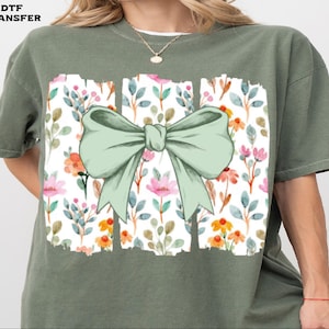 May include: Olive green t-shirt featuring a large, light green bow graphic. The bow is centered over a rectangular design filled with a floral pattern of pink, orange, and blue flowers and leaves. The text "DTF TRANSFER" is in the upper left corner.