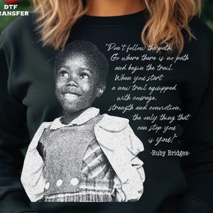 May include: Black sweatshirt featuring a black and white image of Ruby Bridges with a quote. The quote reads, "Don't follow the path. Go where there is no path and begin the trail." The words "DTF TRANSFER" are in the top left corner.