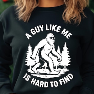 May include: Black sweatshirt with a white graphic of a Bigfoot wearing sunglasses, walking in front of pine trees. The text "A GUY LIKE ME IS HARD TO FIND" is printed above and below the graphic.