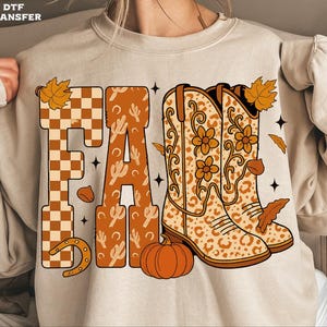 May include: Beige sweatshirt with the word "FALL" in a fall-themed design. The letter "F" is a checkered pattern, the "A" has cactus designs, and the "L"s have cowboy boots and a pumpkin. Autumn leaves and a horseshoe complete the design.