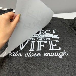 May include: Dark gray t-shirt with white text that reads "I have a freaking awesome wife, that's close enough." A hand is peeling away a transfer sheet, revealing the design. The shirt is on a white surface.