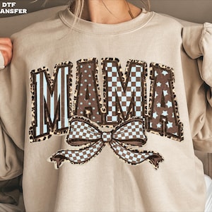 May include: A beige sweatshirt with the word "MAMA" in a decorative design. The letters have a brown leopard print border, with blue and white checkered, polka dot, and star patterns inside. A bow with a checkered pattern is below the word.
