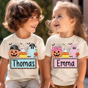 May include: Two cream-colored t-shirts featuring Halloween graphics. One shirt displays a pumpkin, candy corn, and a blue plaid ghost with the name Thomas. The other shirt shows a pumpkin, candy corn, and a pink plaid ghost with the name Emma.
