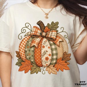 May include: A cream-colored t-shirt featuring a large, central graphic of a pumpkin composed of various fabric-like patterns in shades of orange, green, and white. The pumpkin is adorned with a plaid bow and surrounded by autumn leaves.