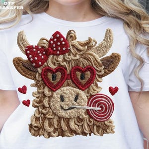 Pode incluir: Camiseta branca com um desenho bordado de uma vaca Highland com &oacute;culos de sol em forma de cora&ccedil;&atilde;o, um la&ccedil;o vermelho e um pirulito. A vaca tem um rosto sorridente e &eacute; cercada por pequenos cora&ccedil;&otilde;es vermelhos.