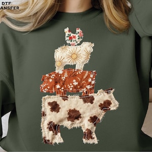 May include: Dark green sweatshirt featuring a stack of farm animals: a cow, a pig, a sheep, and a chicken. The animals are made of fabric with floral and sunburst patterns. The text "DTF TRANSFER" is in the top left corner.