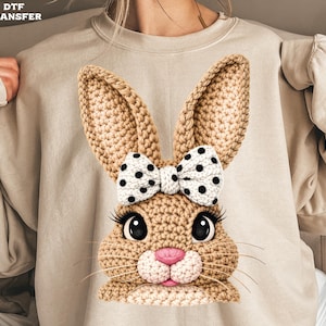 May include: A beige sweatshirt featuring a detailed, crocheted bunny face. The bunny has large, expressive eyes, long eyelashes, a pink nose, and a white bow with black polka dots. The text "DTF TRANSFER" is in the upper left corner.
