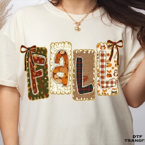 May include: A cream-colored t-shirt featuring the word FALL in large, decorative letters. Each letter is contained within a rectangular patch with distinct patterns including pumpkins, plaid, and foxes, accented by small bows on the F and L.