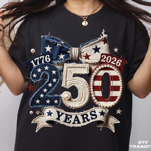 May include: Black t-shirt with a patriotic design. Features a bow and the numbers "250" and "0" with the years "1776" and "2026". The design includes stars and stripes, and the words "YEARS".