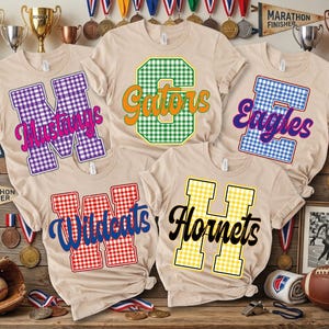 May include: Five beige t-shirts featuring large gingham-patterned letters with team names overlaid in script. The designs include a purple M with Mustangs, a green C with Gators, a blue E with Eagles, a red W with Wildcats, and a yellow H with Hornets.