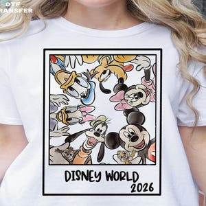 May include: White t-shirt featuring a framed graphic with Disney characters including Mickey Mouse, Minnie Mouse, Donald Duck, and Goofy. The graphic reads "DISNEY WORLD 2026". The shirt has a DTF TRANSFER logo in the top left corner.