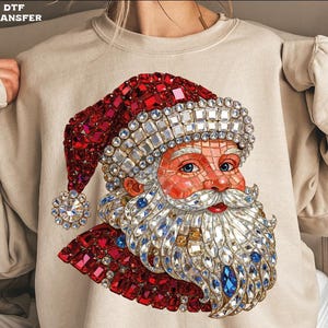 May include: A beige sweatshirt featuring a detailed Santa Claus design. The Santa Claus graphic is made of red, white, and blue jewel-like elements, creating a sparkling effect. The text "DTF TRANSFER" is in the upper left corner.