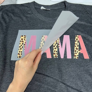 May include: Dark gray t-shirt with the word "MAMA" in pink and leopard print lettering. The shirt is being prepared with a heat transfer vinyl design. Other shirts are visible in the background.